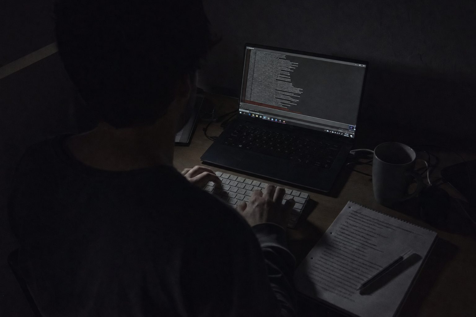 A desk at one in the morning, lit only by the monitor. Hands on a keyboard, a terminal full of scrolled output, and a notepad with every line crossed out.